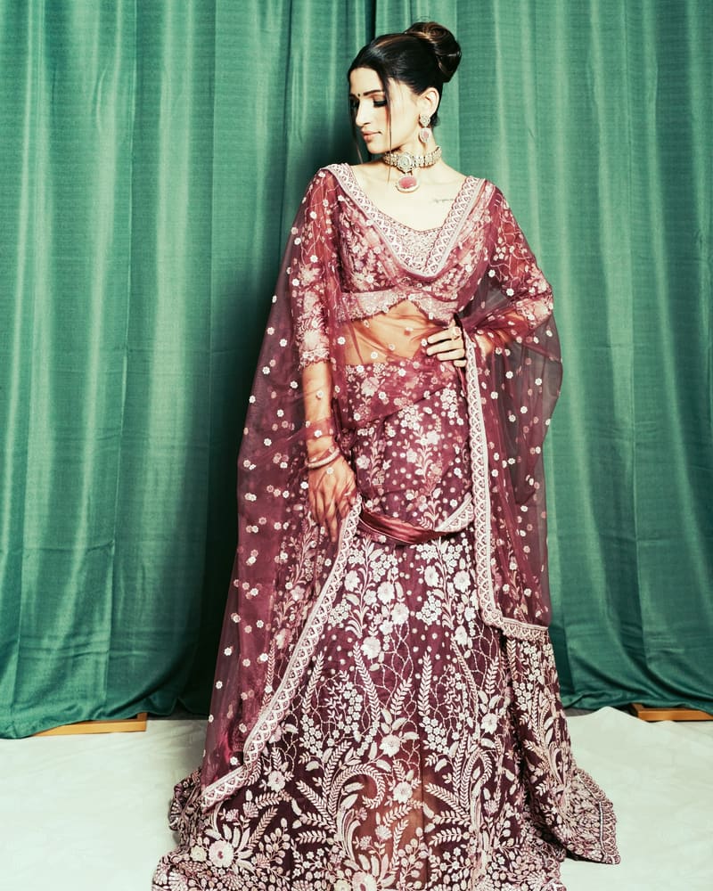 Model in a wine lehenga with tonal sequin embroidery and a sheer bordered dupatta, standing in front of an emerald curtain