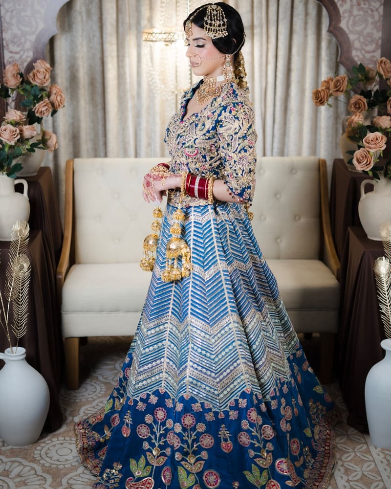 Bride in a blue chevron lehenga standing beside a cream sofa with dried flowers