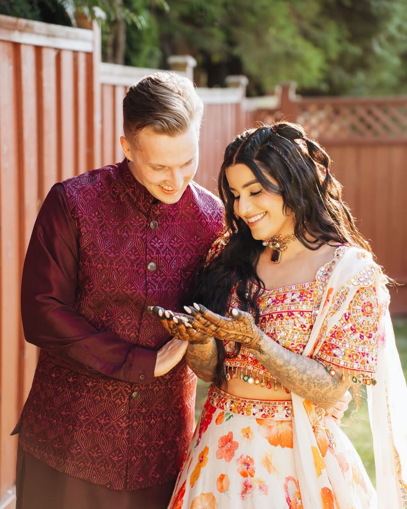 Couple at their mehendi, she in a floral lehenga showing her henna to him in a maroon kurta