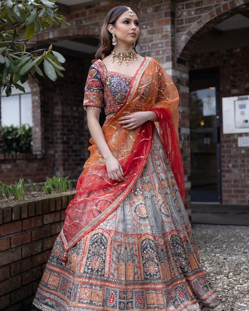 Woman in a multicolour lehenga with an orange dupatta standing outside a brick building