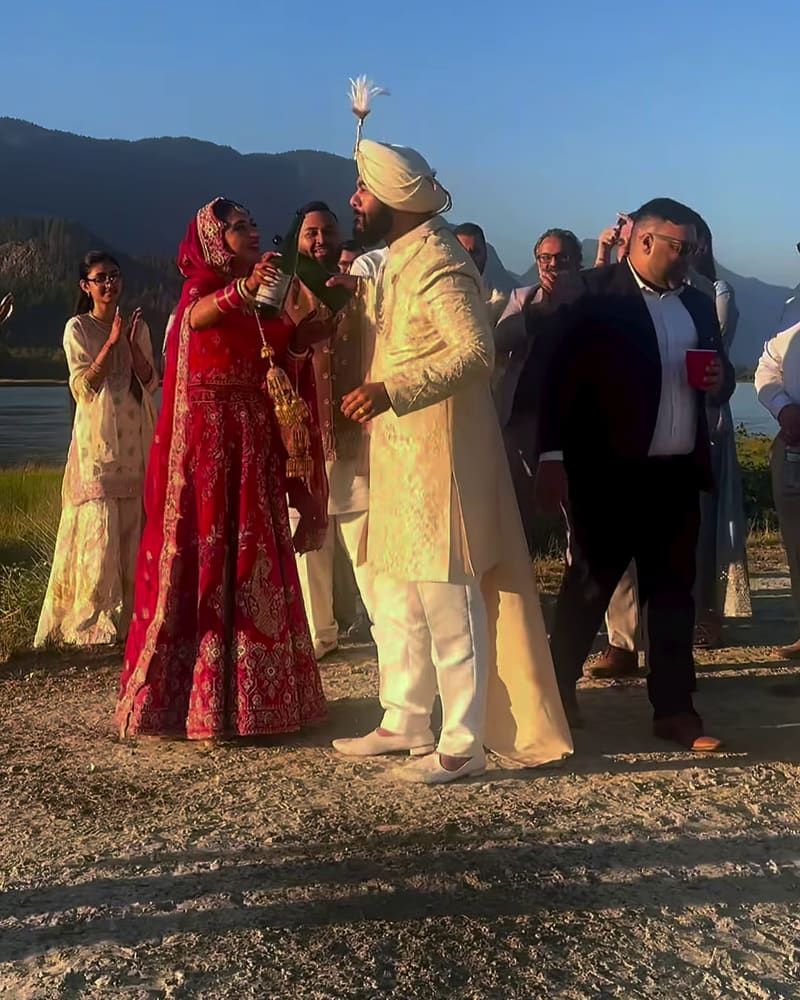 Bride in red and groom in ivory walking together by the water with family behind them